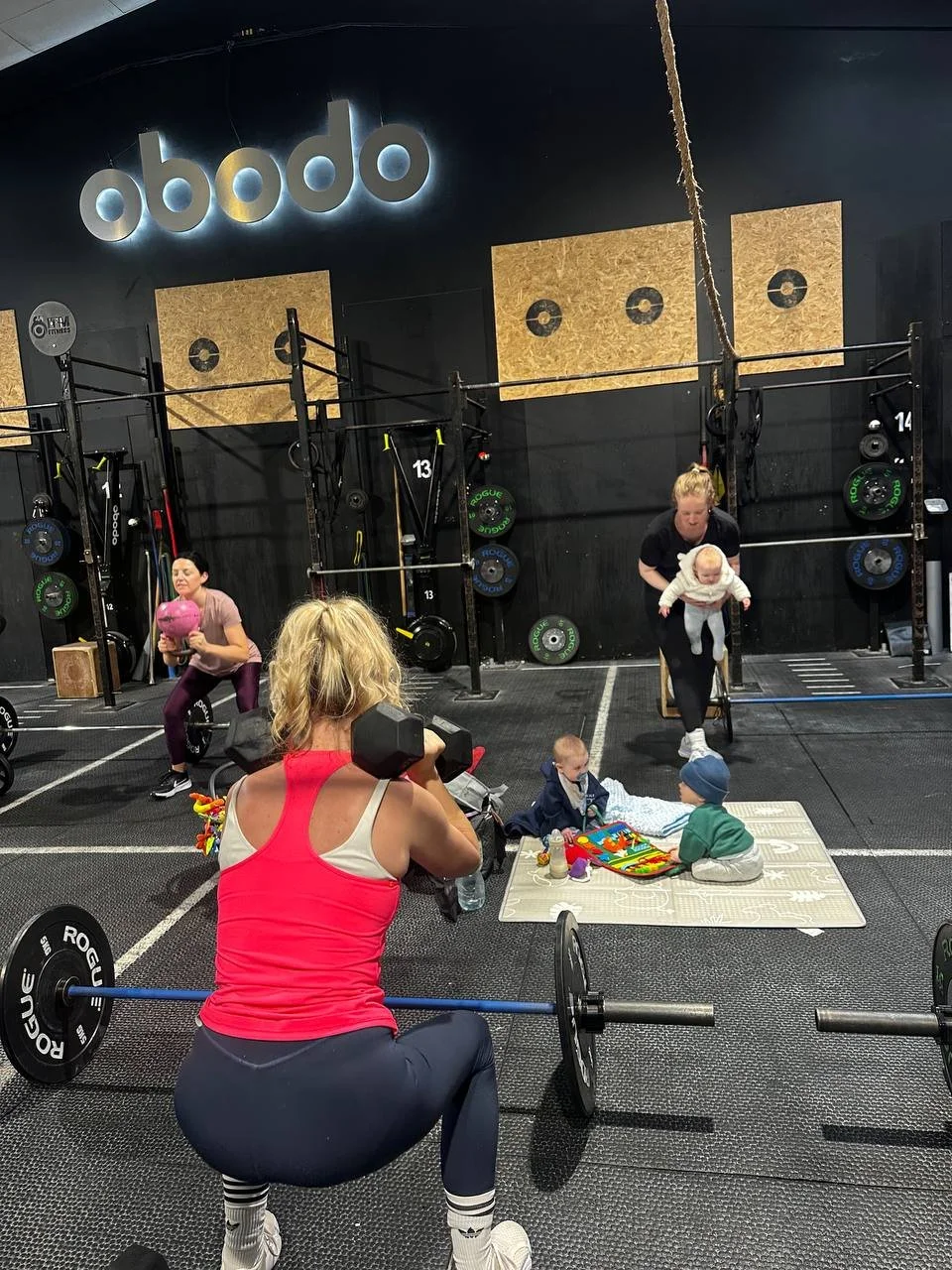 People working out at a gym with children and a woman taking a photo. The gym has weights and workout equipment, and the scene includes a group of children playing on a mat.