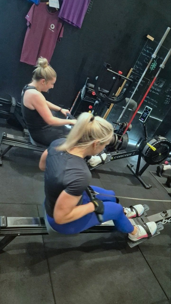 Two women using rowing machines in a gym, with black walls, exercise equipment, and a chalkboard in the background.