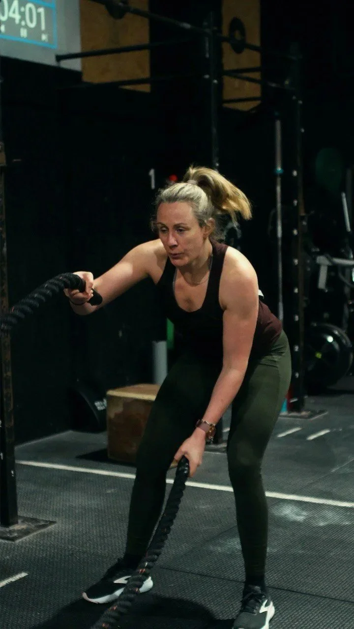 Woman in black workout attire pulling rope in gym with black floors and fitness equipment.
