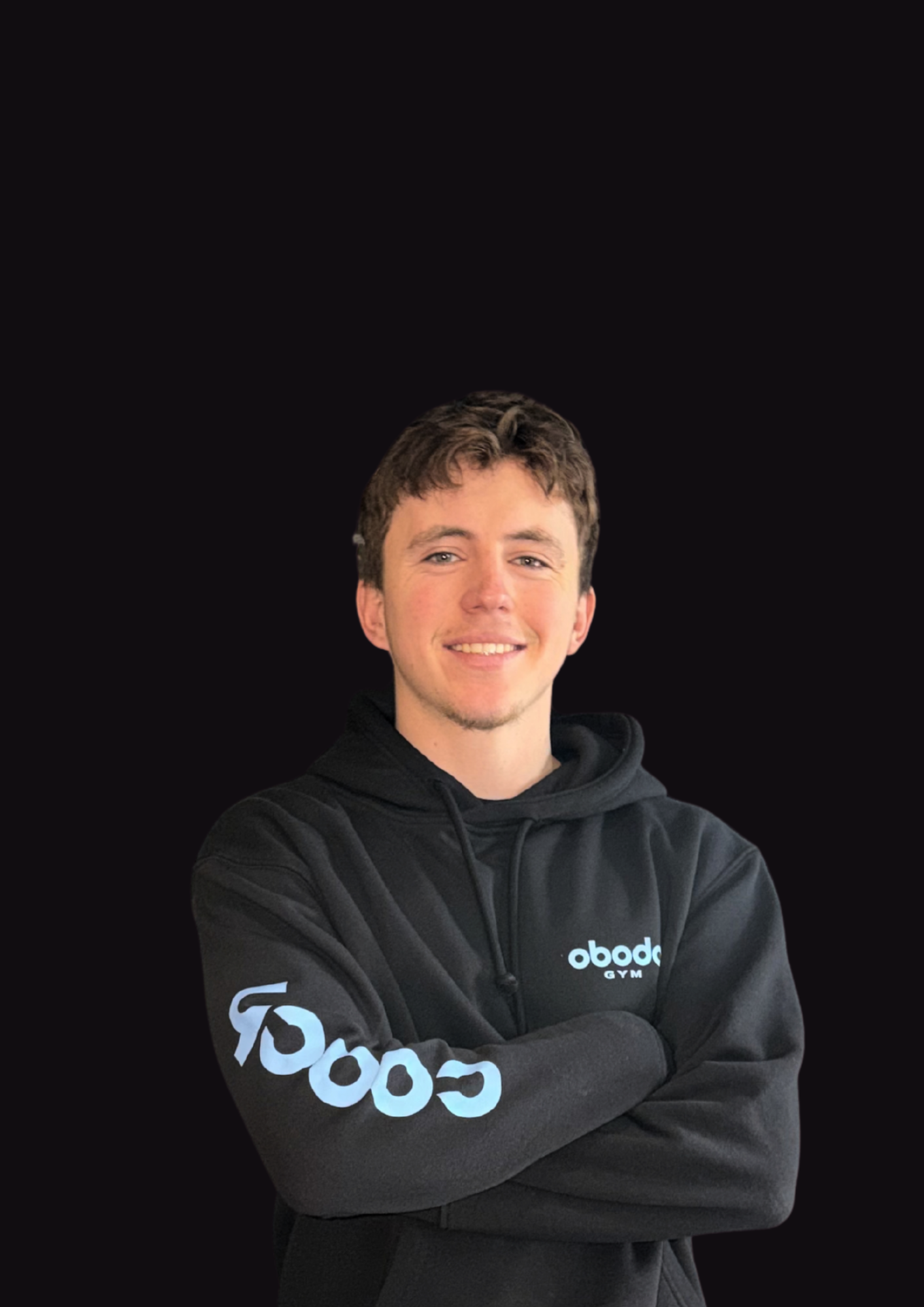 A young man with short brown hair and a light beard, smiling with arms crossed, wearing a black hoodie with 'Obode Gym' logos on the chest and sleeve, standing against a black background.