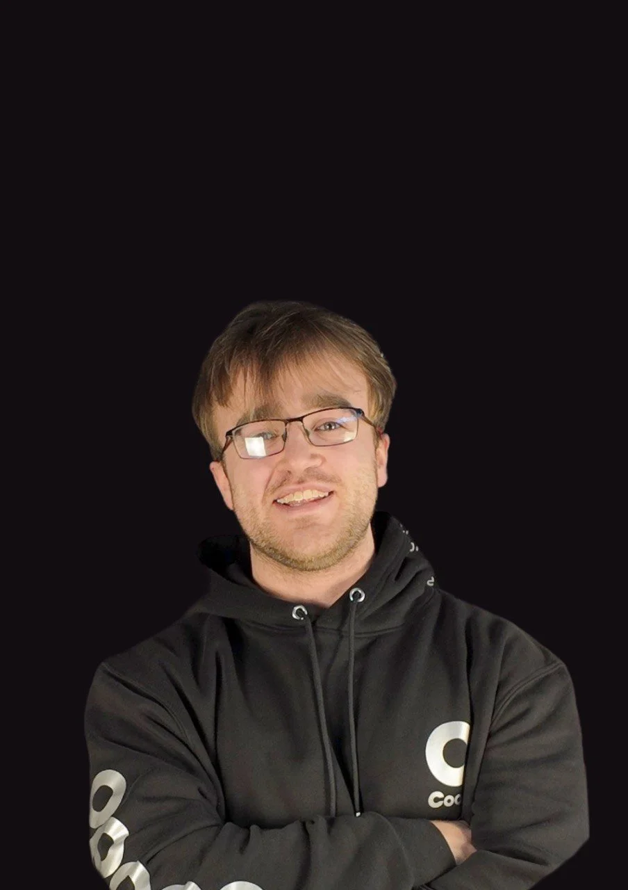 A man with brown hair, glasses, and a beard wearing a black hoodie with white text on the sleeves and chest, smiling with arms crossed against a plain black background.