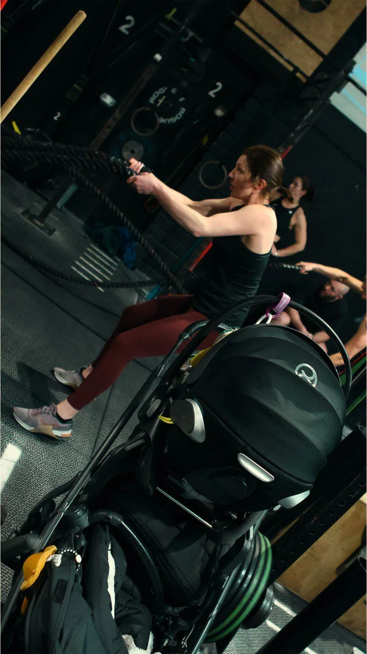 A woman exercising with battle ropes in a gym, with a stroller in the foreground.