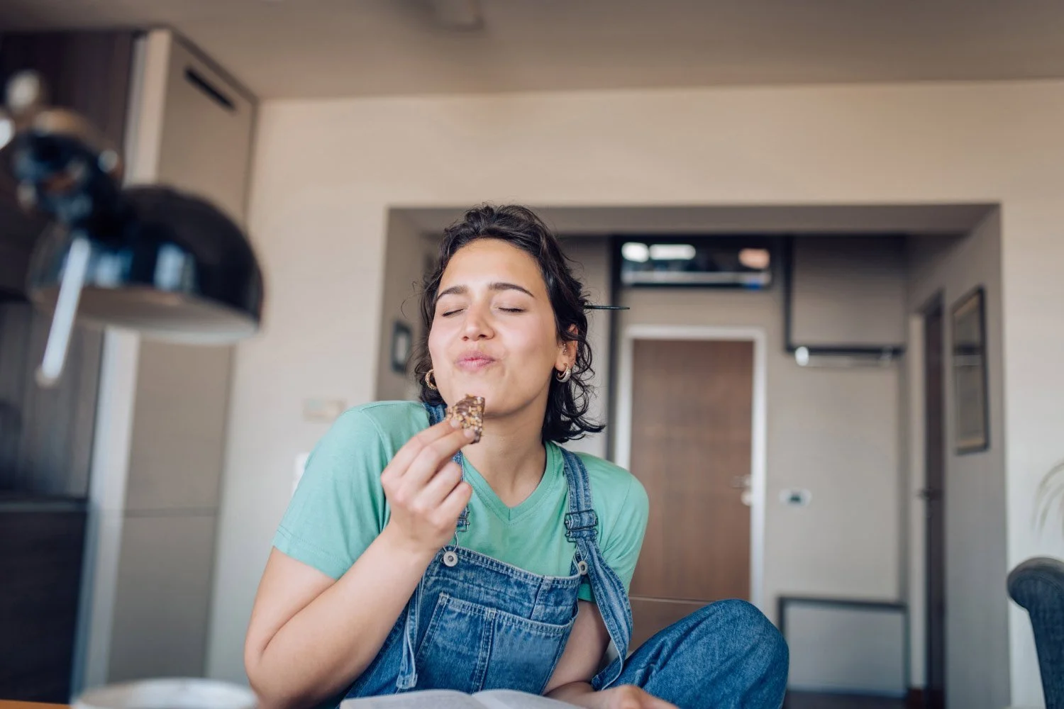 Young woman with short dark curly hair wearing a green t-shirt and denim overalls, enjoying a chocolate cookie with eyes closed in a cozy indoor space.