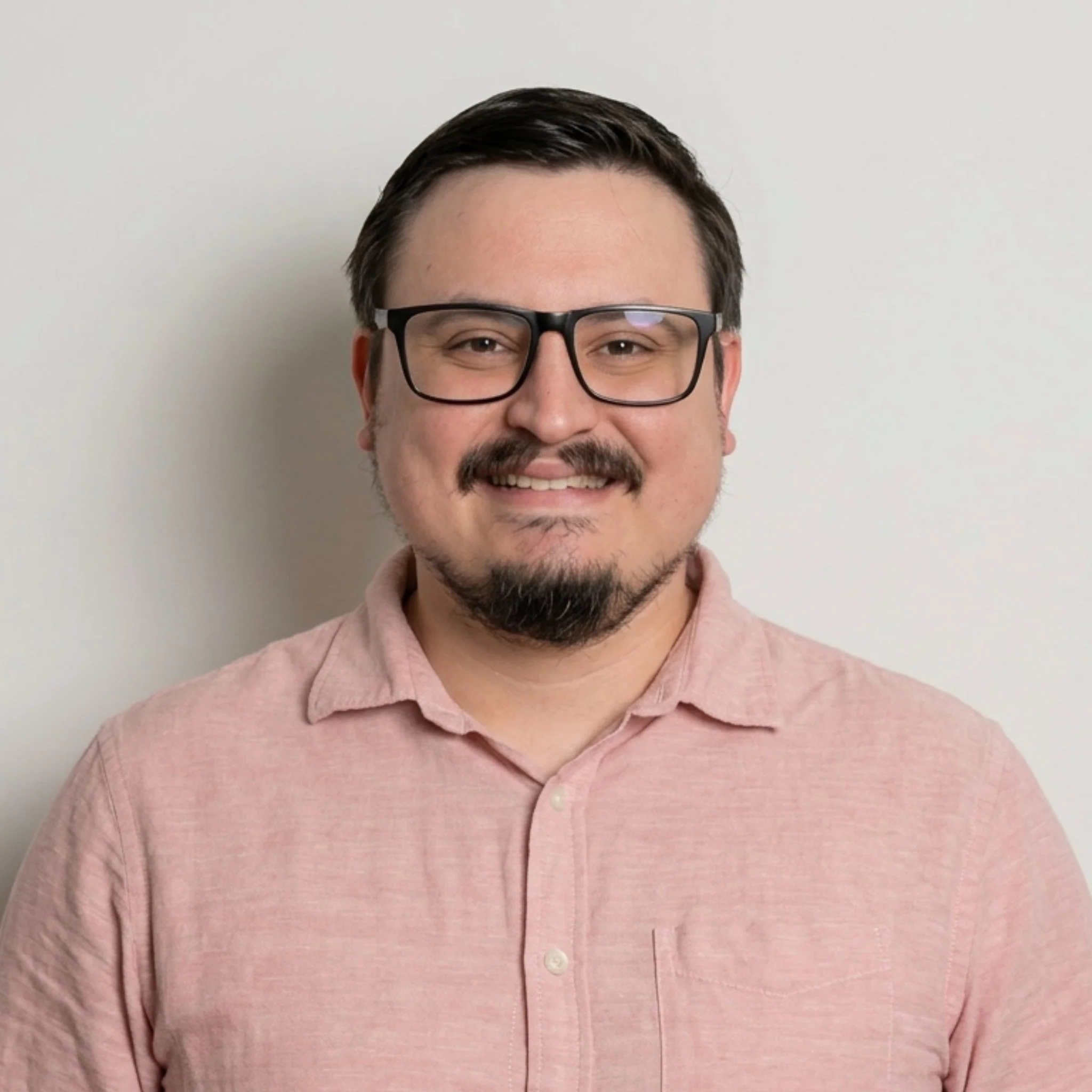 Close-up of a man's face with glasses, smiling, wearing a light pink shirt, against a plain light gray background.
