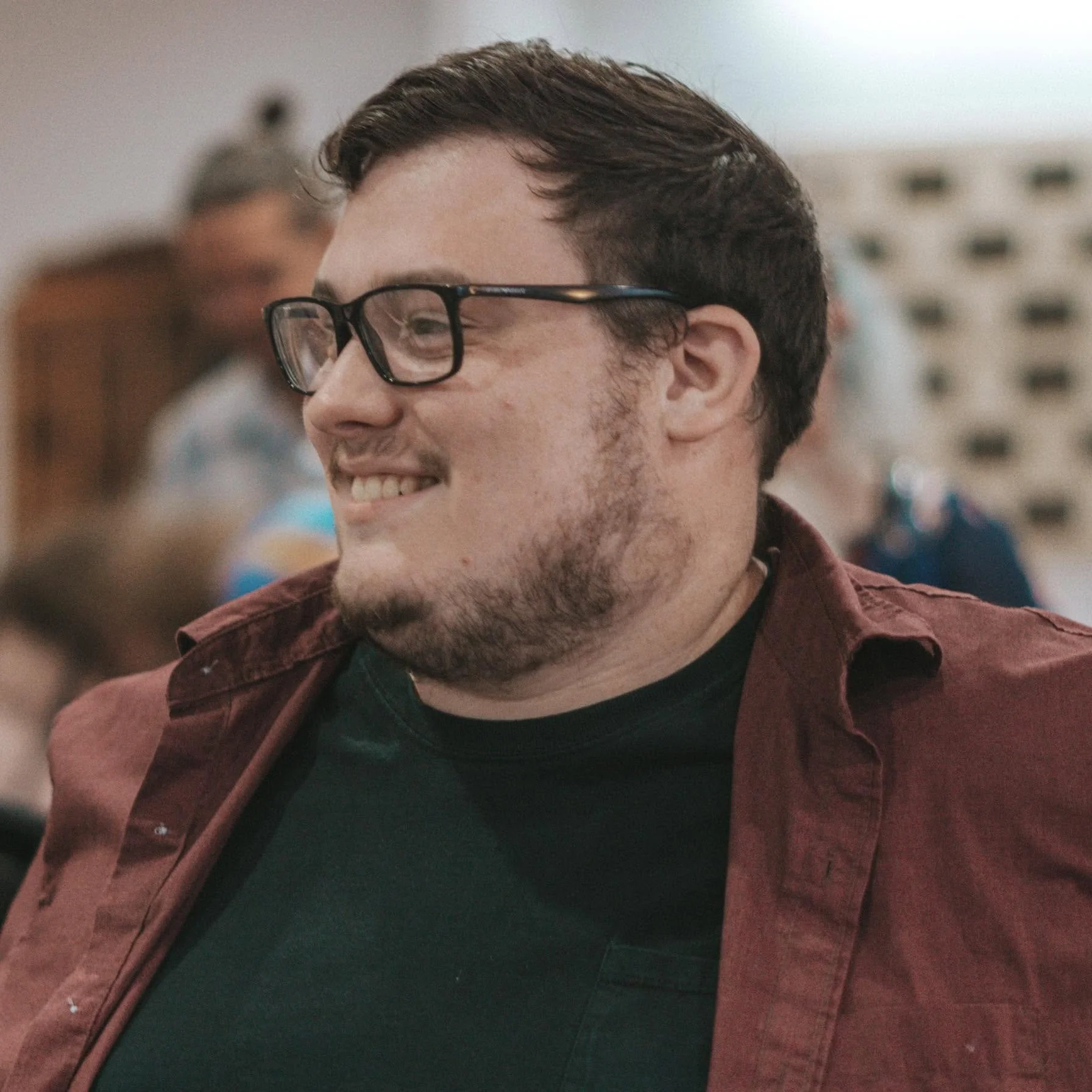 Smiling man with glasses and short brown hair, wearing a dark shirt and a maroon jacket, in a crowded indoor setting.