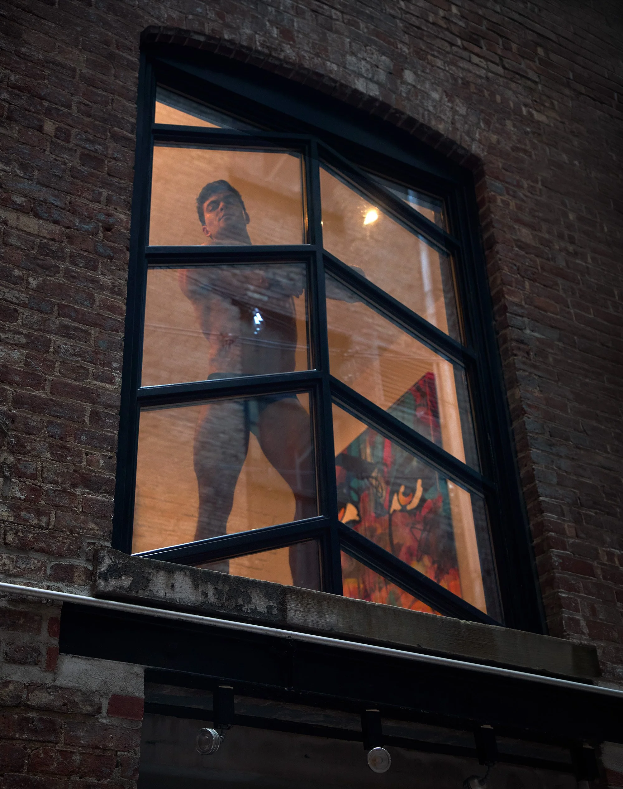 A man with styled hair looking out of a modern black-framed glass window in a brick building at dusk.