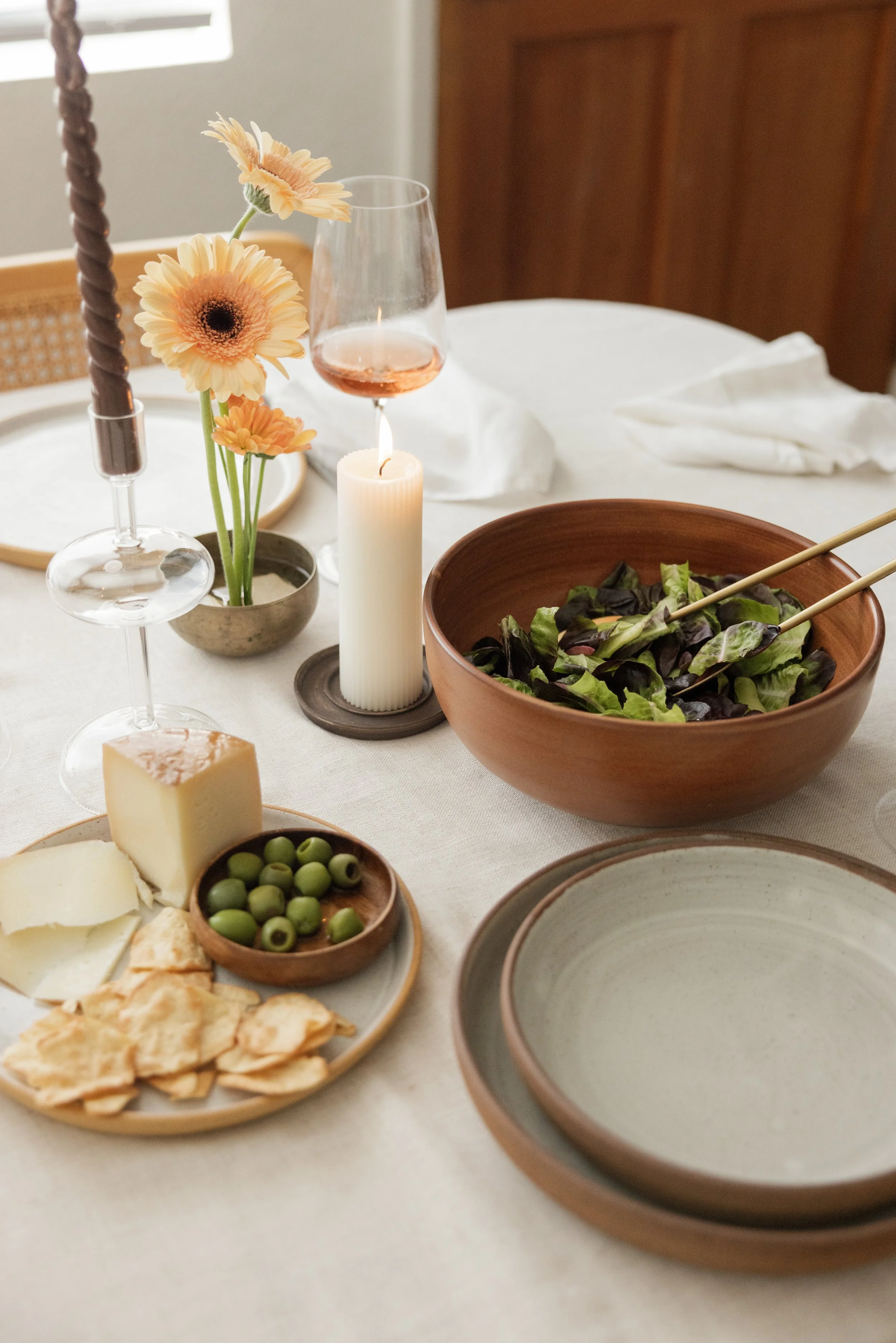 A dinner table set with a salad bowl, wine glass, candle, cheese, olives, and bread, decorated with a small flower arrangement.