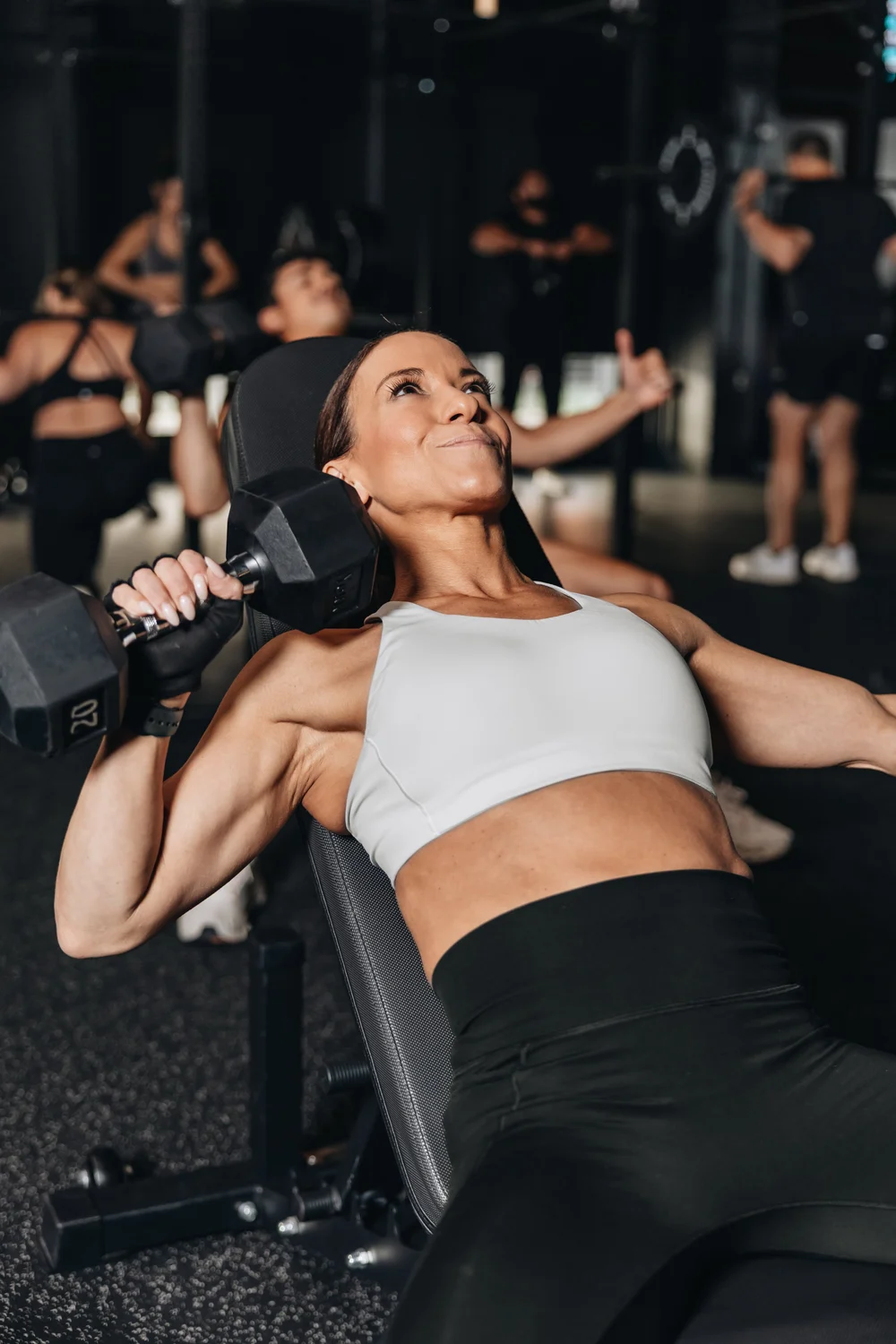 A woman on an incline bench mid-press, head back and arms loaded, clients training in the background