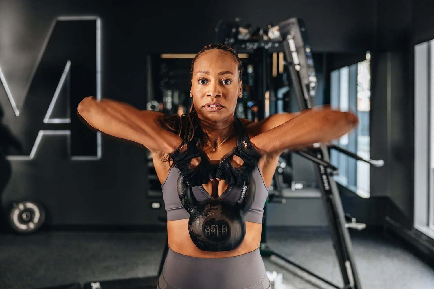A woman raising a kettlebell to shoulder height with both hands, looking straight ahead, the TRAIN Moment M on the wall behind her