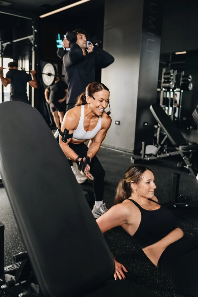 Two women either side of an incline bench, one seated on the floor and one standing behind it with a dumbbell