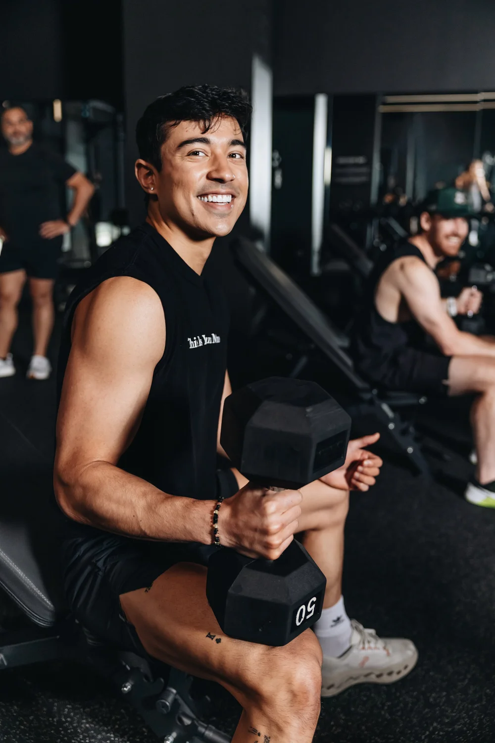 A man seated on a bench holding a dumbbell, grinning broadly, other clients training behind him