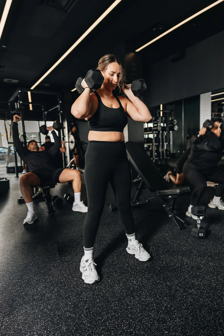 A woman standing with a dumbbell racked at each shoulder, other clients training behind her