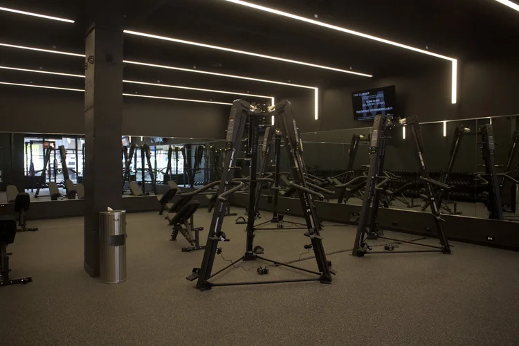 Three VersaClimbers on the training floor in front of the mirrored wall, benches and dumbbell racks behind them and a screen showing the session