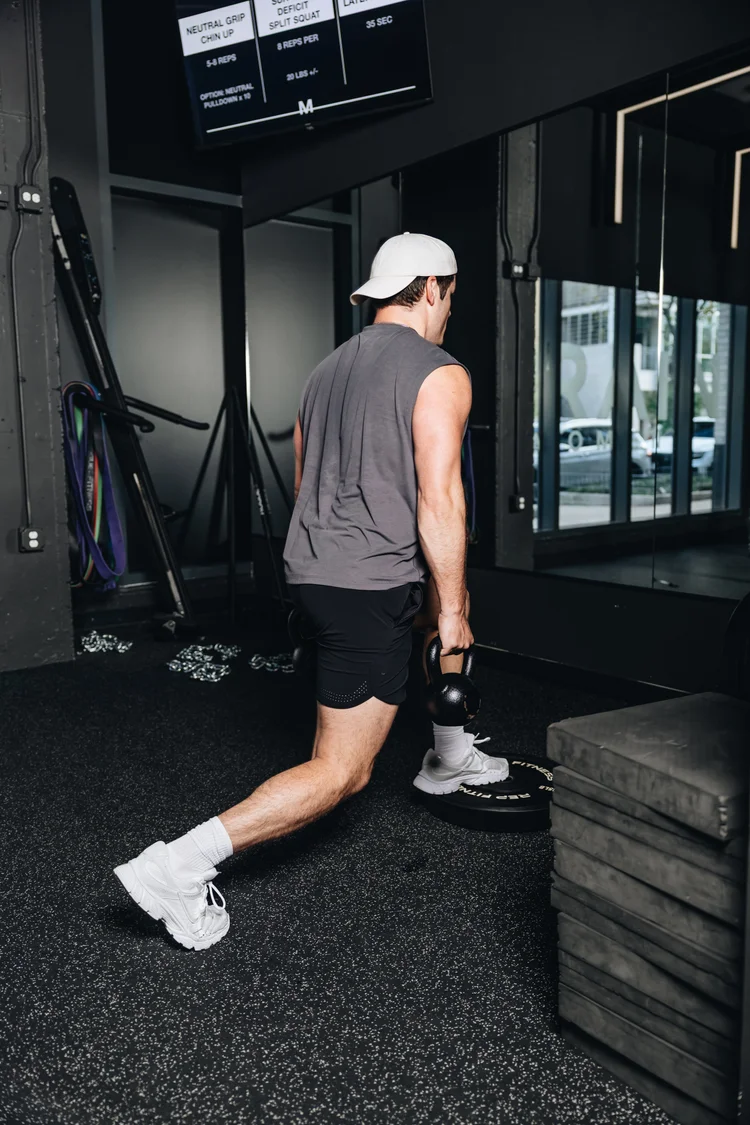 A man carrying a kettlebell across the floor toward a set of boxes, daylight through the studio windows behind him