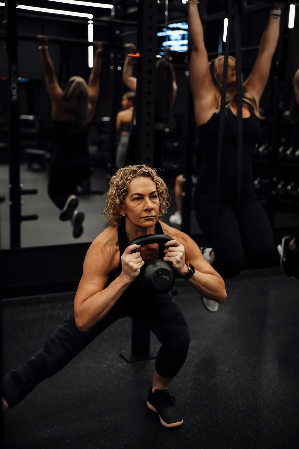 A woman holding a dumbbell at her chest in a deep squat, others training behind her