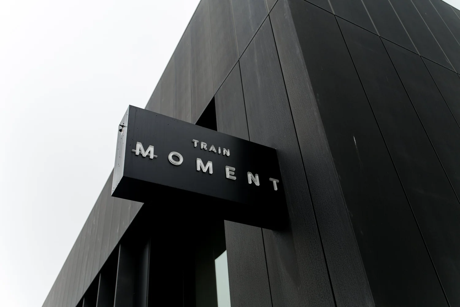 The studio's black metal-clad corner from below, the TRAIN MOMENT blade sign mounted on it, the glass tower rising above and behind