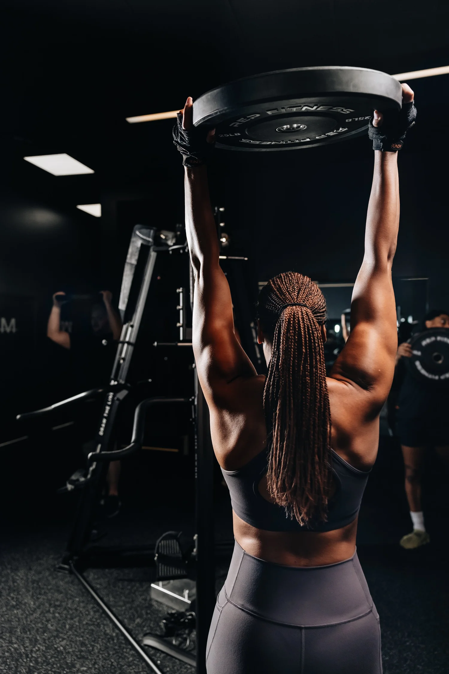 A member pressing a weight plate overhead at TRAIN Moment River North, the TRAIN Moment mark on the wall behind her