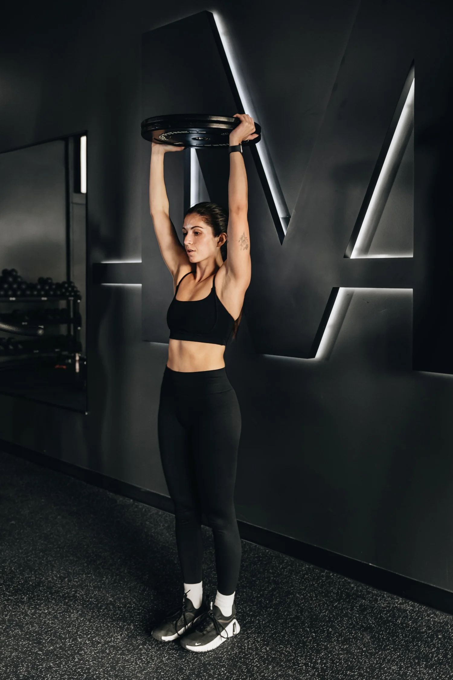 A woman pressing a weight plate overhead with both hands, standing in front of the illuminated TRAIN Moment M on the studio wall