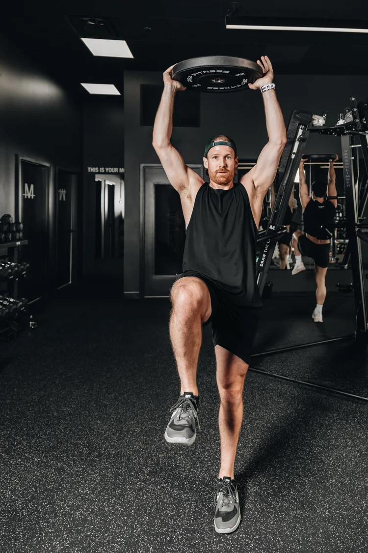 A man striding forward across the floor holding a weight plate locked out overhead