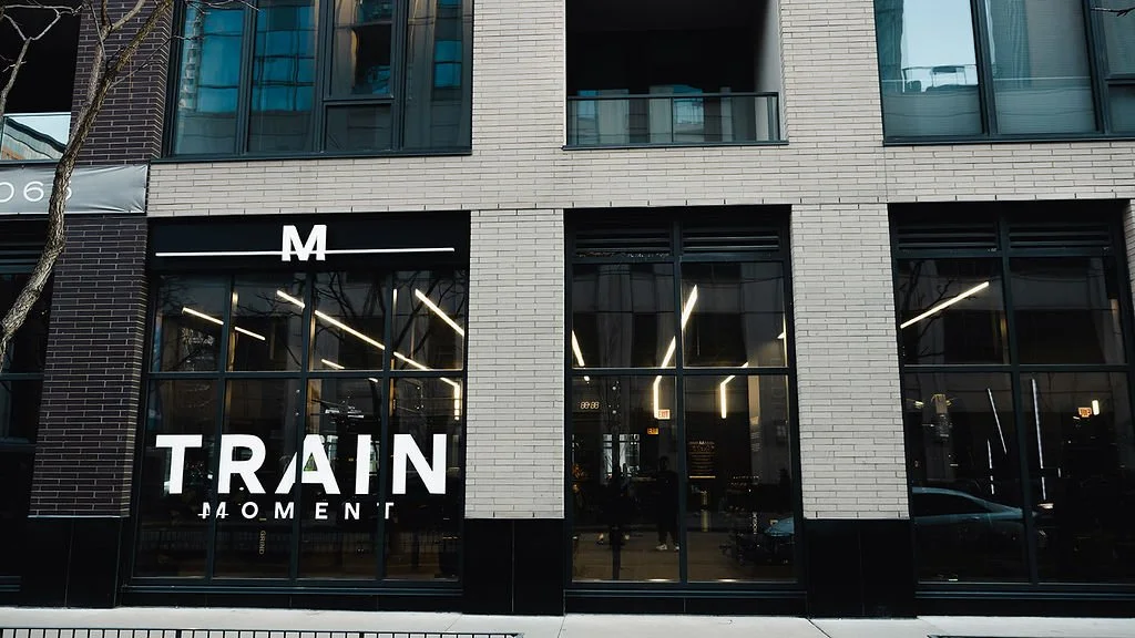 The studio storefront from across the street, the M mark and TRAIN MOMENT across the black window band, the entrance between two brick piers