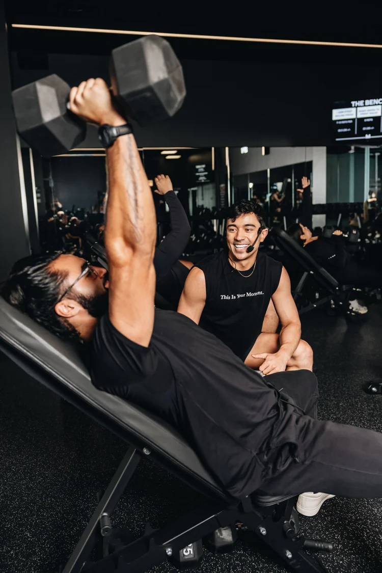 A man pressing a dumbbell overhead on an incline bench while a coach sits alongside him watching the set