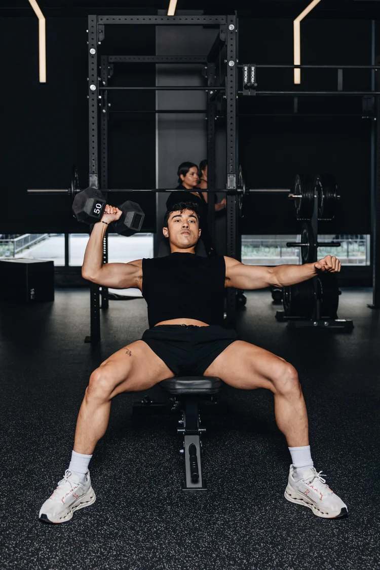 A person seated on a bench pressing two dumbbells with arms wide, a squat rack behind them