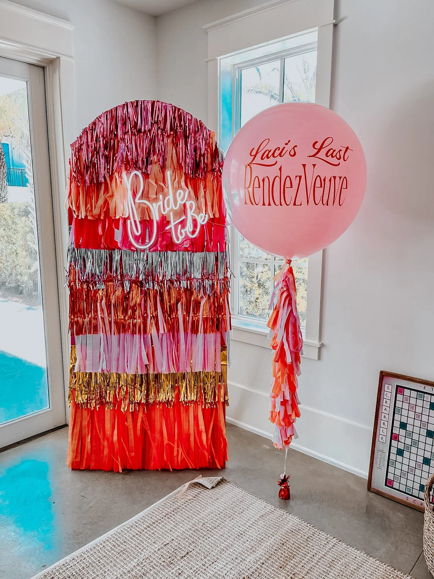 Colorful fringe photo backdrop with a custom giant pink balloon reading “Laci’s Last RendezVous” and a “Bride to Be” neon sign styled for a 30A bachelorette celebration.
