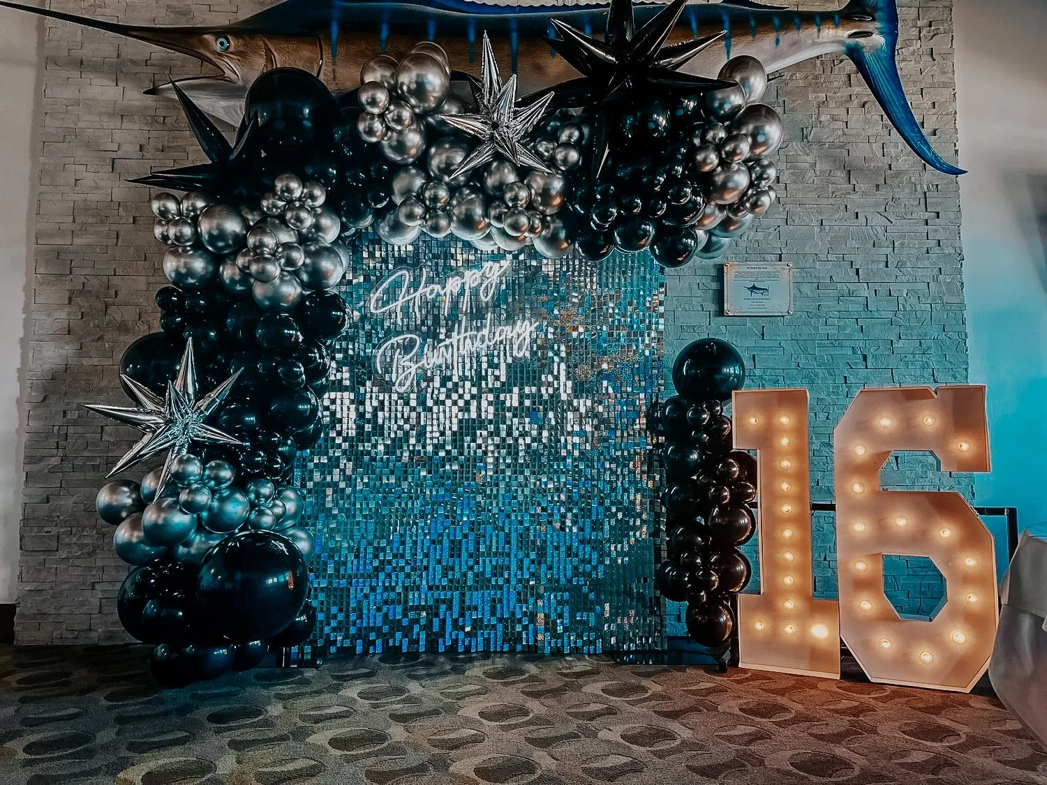 Black, silver, and blue balloon décor with stars, a shimmer wall backdrop, neon “Happy Birthday” sign, and an illuminated “16” for a 30A birthday celebration.