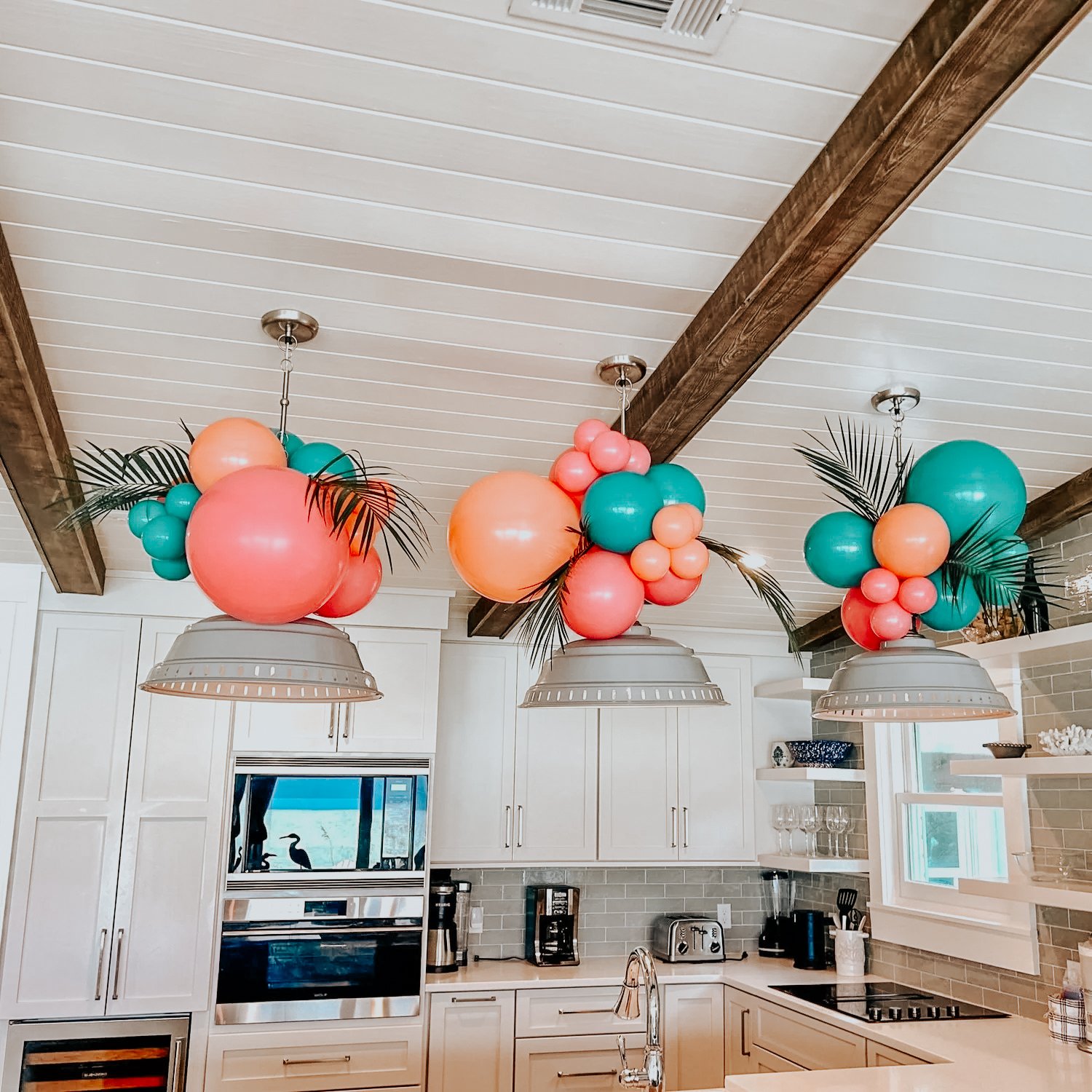 Pink, teal, and peach balloon clusters with palm leaves hanging from kitchen pendant lights for a 30A indoor celebration.