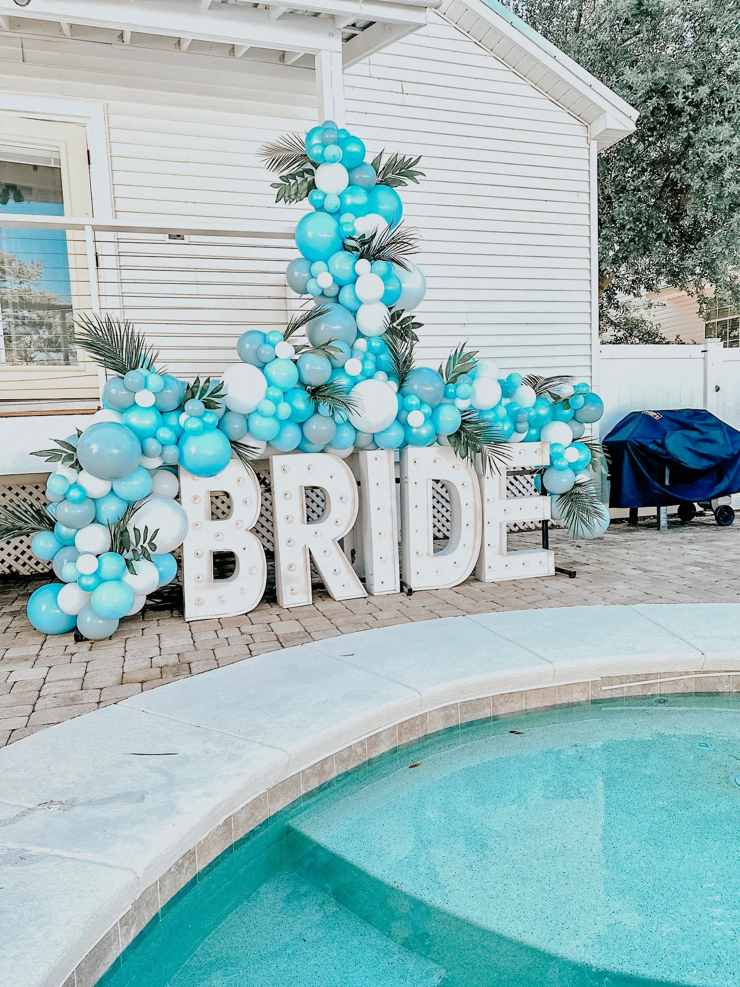 Blue, white, and gray balloon décor with greenery and a “BRIDE” sign styled for an outdoor 30A bridal celebration near a pool.