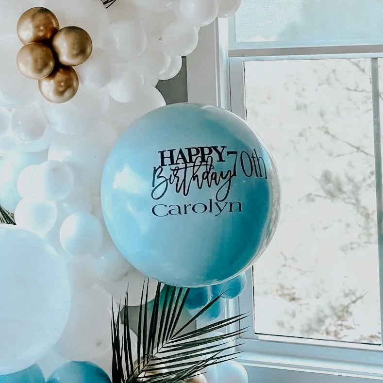 Custom giant blue balloon with “Happy 70th Birthday Carolyn” lettering surrounded by white and gold balloon garland for a 30A milestone celebration.