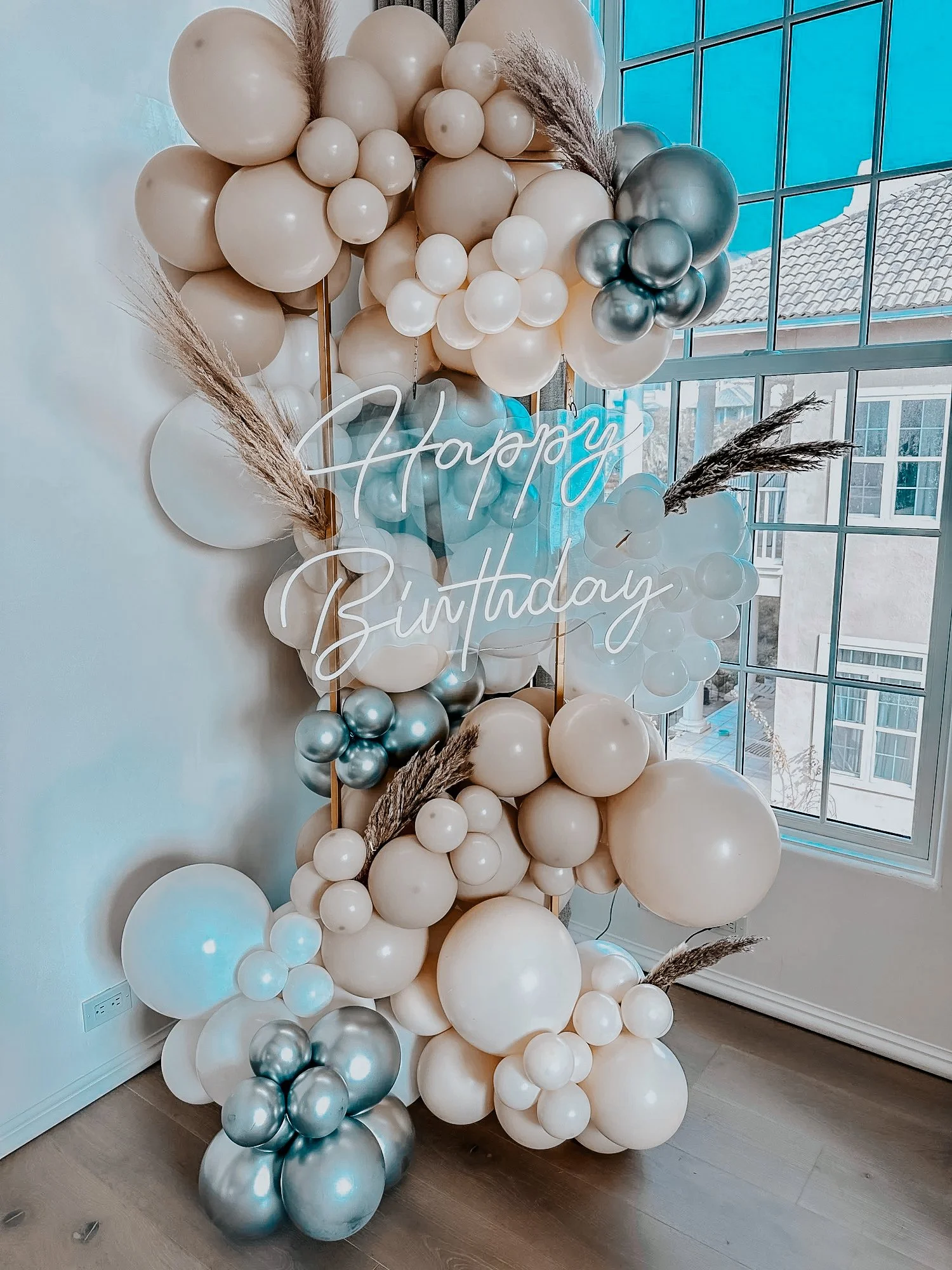Cream, white, and metallic silver balloon garland décor with dried grass accents and a “Happy Birthday” neon sign styled for a 30A birthday celebration.