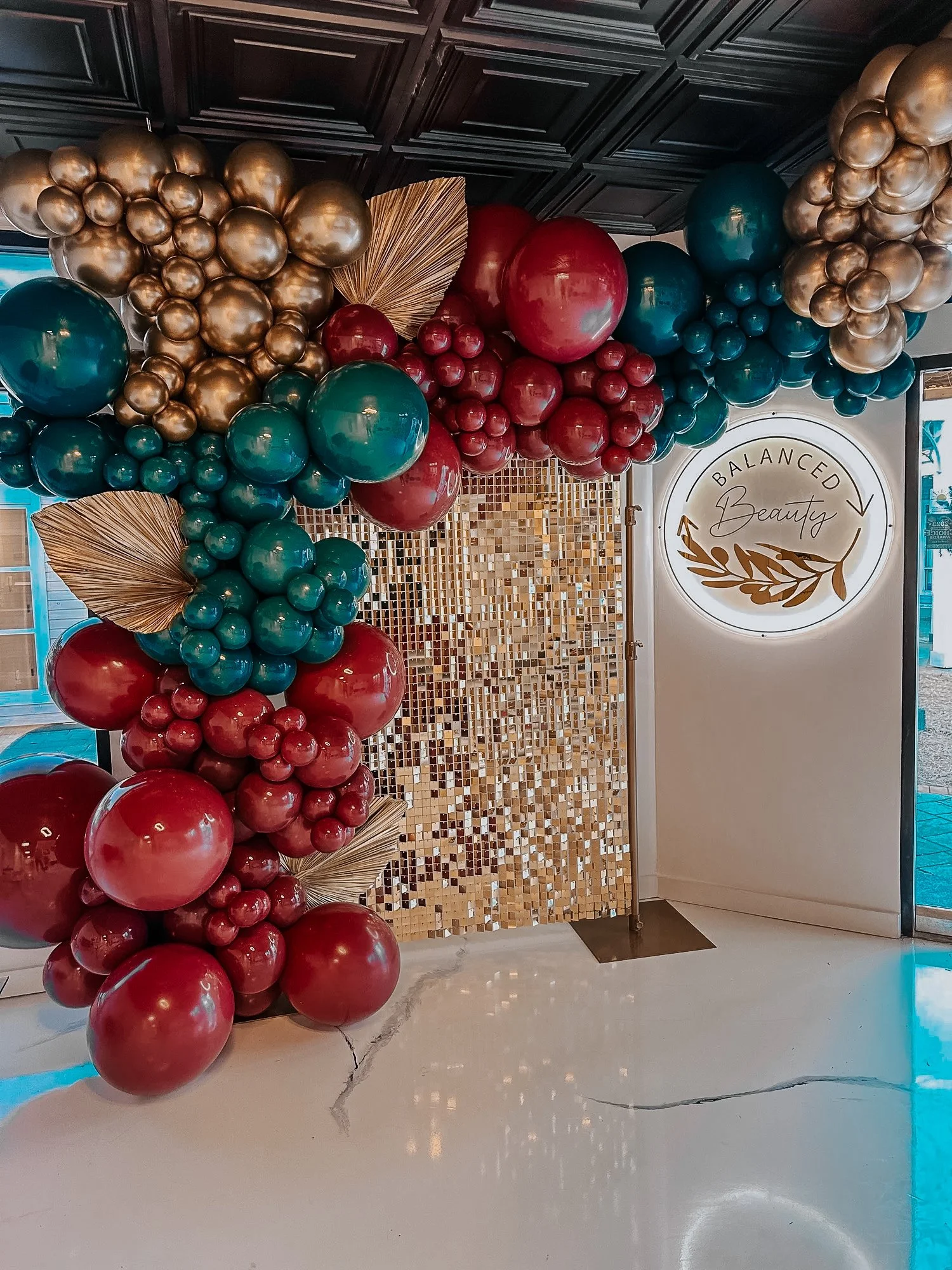 Gold, green, and burgundy balloon arch styled for a 30A business event in front of the Balanced Beauty illuminated wall.