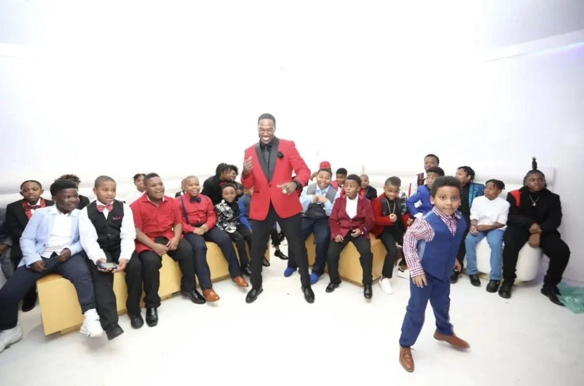 A man in a red blazer and black pants dancing in front of a group of children in a bright room.