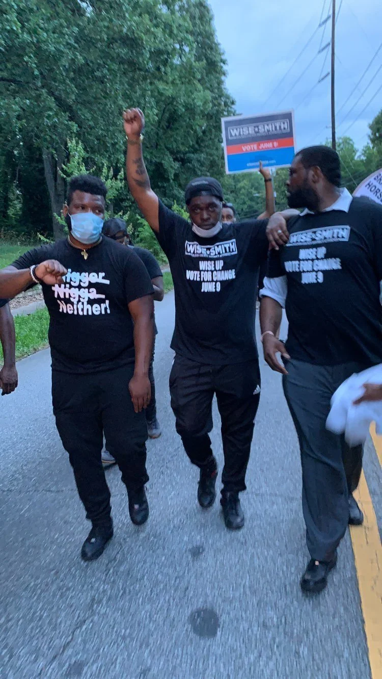 Group of Black people participating in a political march or rally outdoors during daytime, wearing black T-shirts with campaign messages, one person raising their fist in the air, some wearing masks, with campaign signs in the background supporting candidate Wis Smith.