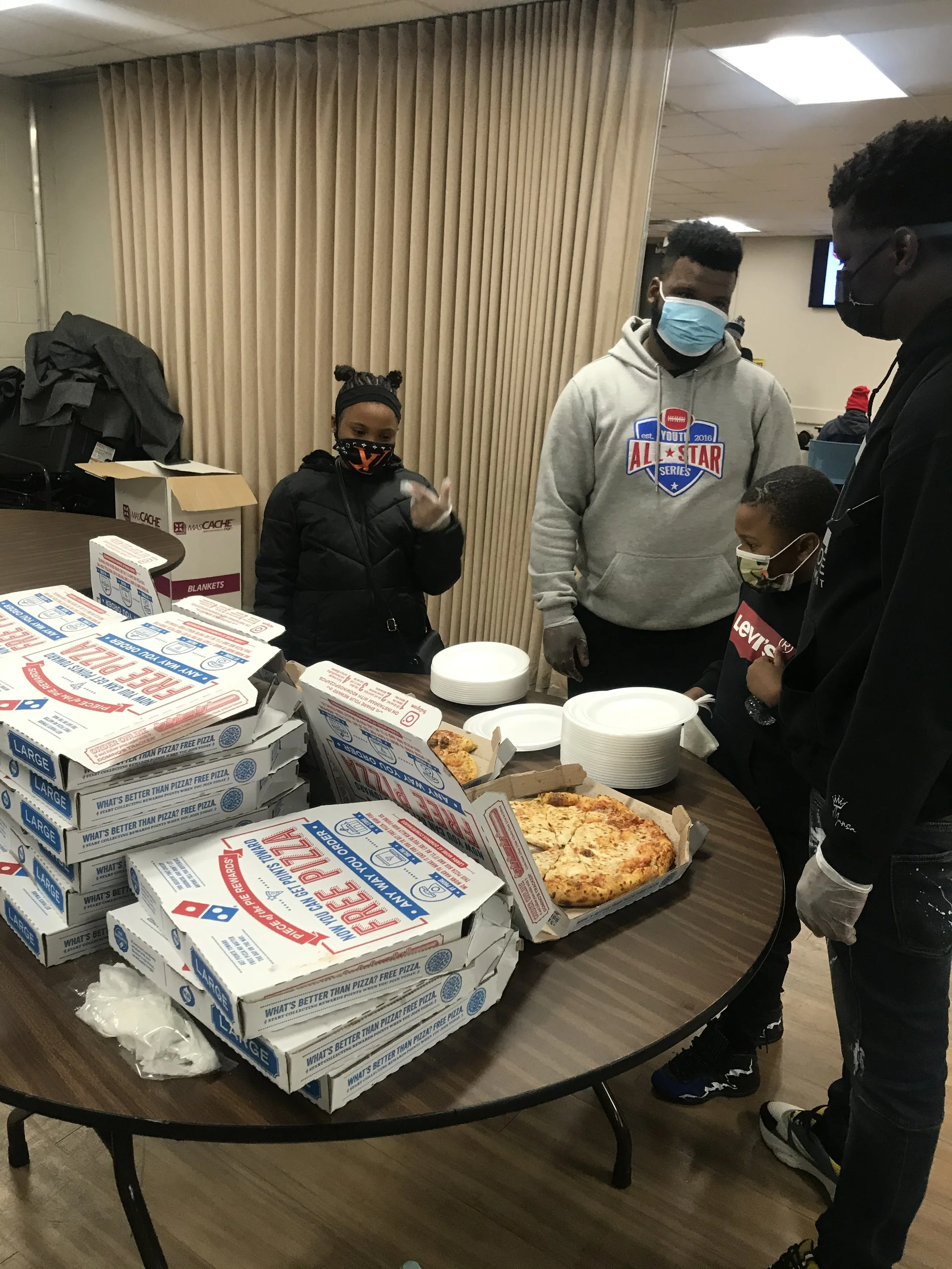 Four people wearing masks standing around a table with pizza boxes, plates, and pizza slices.
