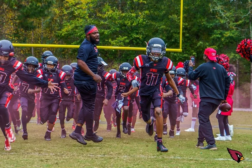 A youth American football team wearing black jerseys with red and white accents running onto the field while a coach and a person with a phone record the event.