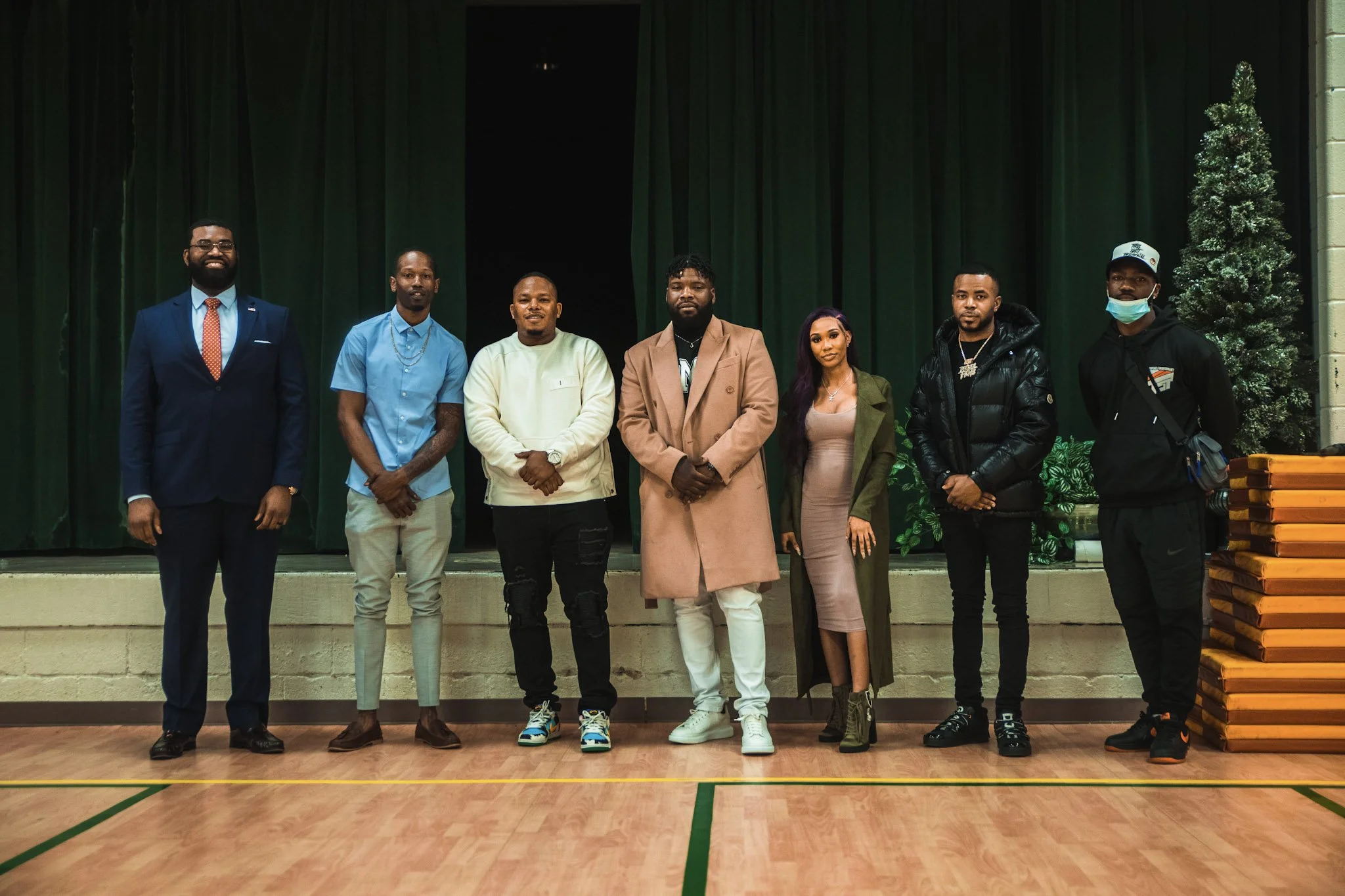 Group of seven diverse people standing in a line on a wooden floor in front of a stage with green curtains, some wearing formal and casual clothing, one person wearing a face mask, with a decorated Christmas tree on the right.