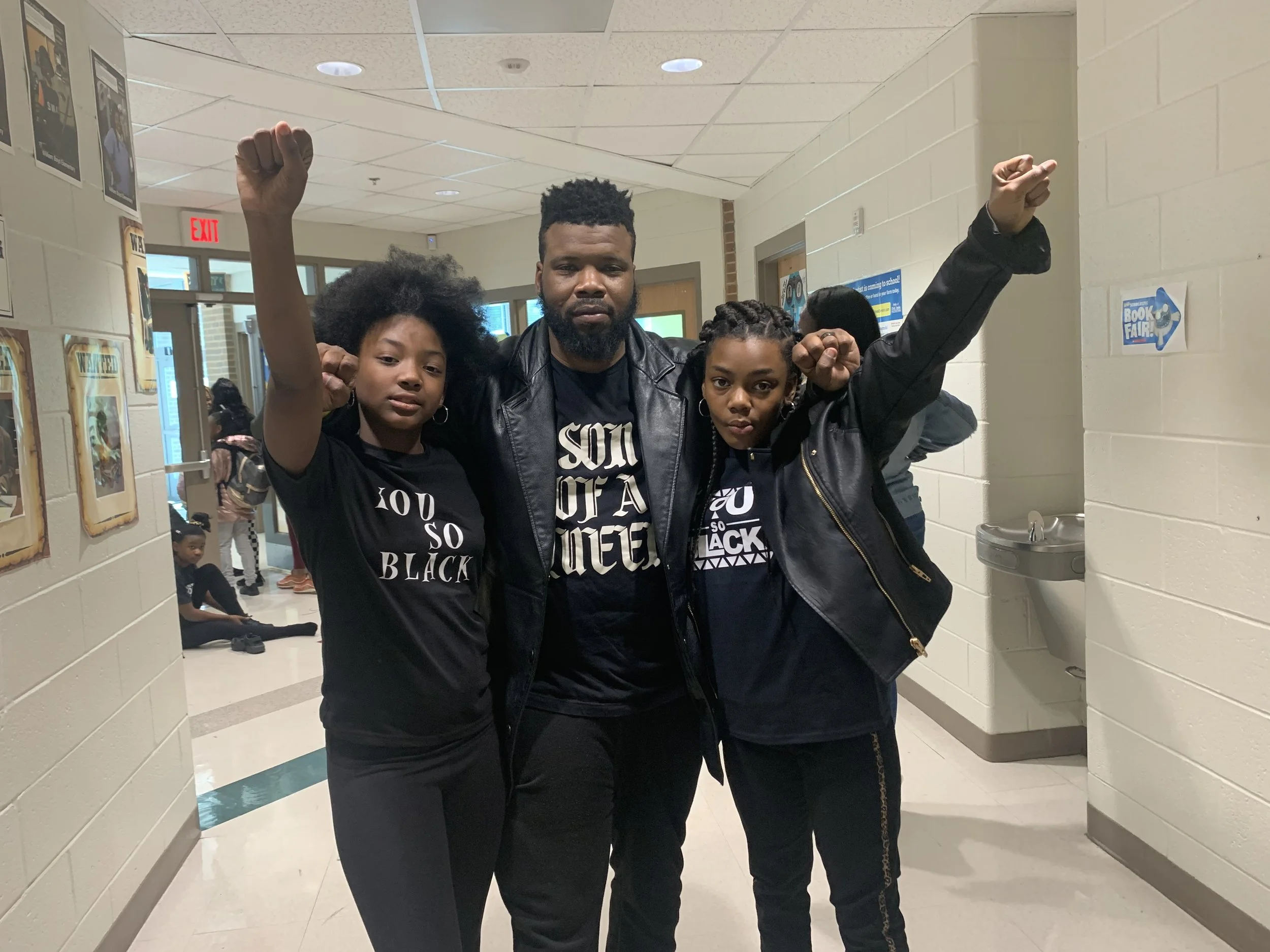 Three individuals standing together in a school hallway, raising their fists in a gesture of solidarity. Two young women and a man, all wearing black clothing with messages supporting Black Lives Matter, are posing confidently.