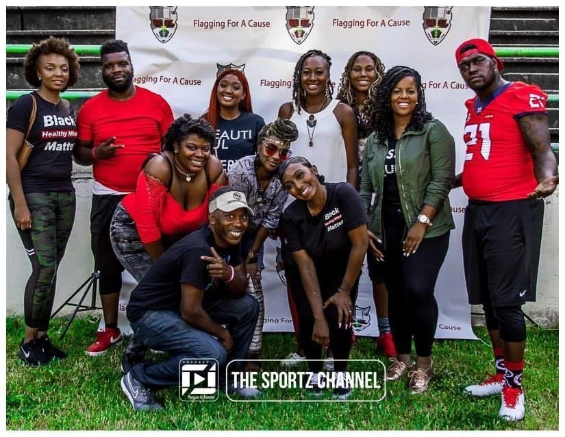 Group of ten smiling people, both men and women, posing outdoors in front of a banner with the text "Flagging For A Cause." The group appears to be at a sports or community event, with some wearing casual and sportswear.