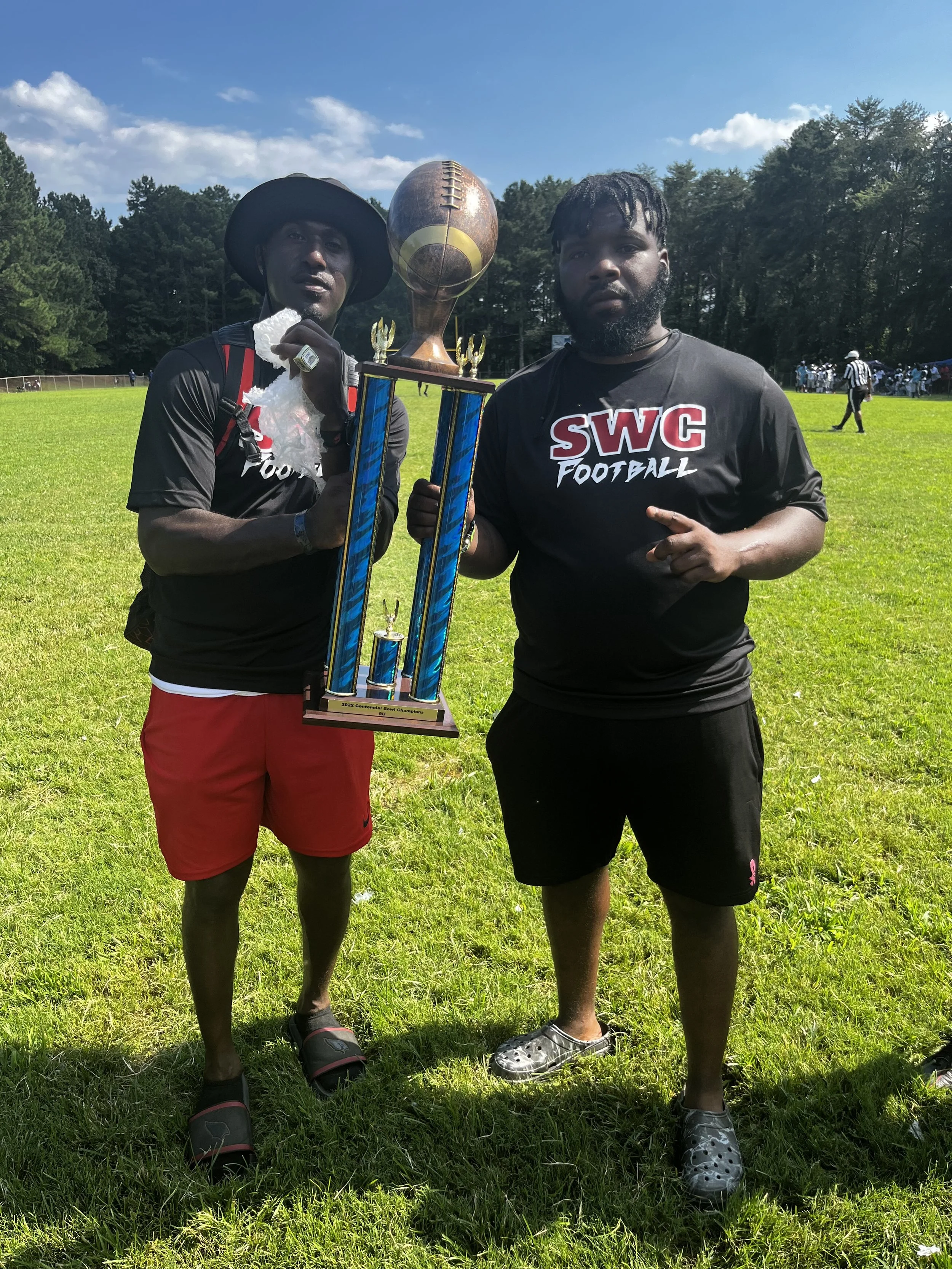 Two men standing on a grassy field holding a tall football trophy. One man is wearing a black SWC football shirt, black shorts, and cleats. The other man is wearing a black hat, black shirt, red shorts, and slides. In the background, there are trees and a football game happening.