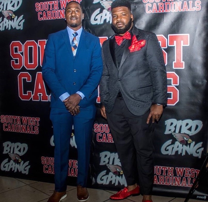 Two men in formal attire standing in front of a backdrop with the text 'South West Cardinals' and 'Bird Gang'. One man is wearing a blue suit with a blue and orange striped tie, and the other man is wearing a black tuxedo with a red bow tie and red shoes.