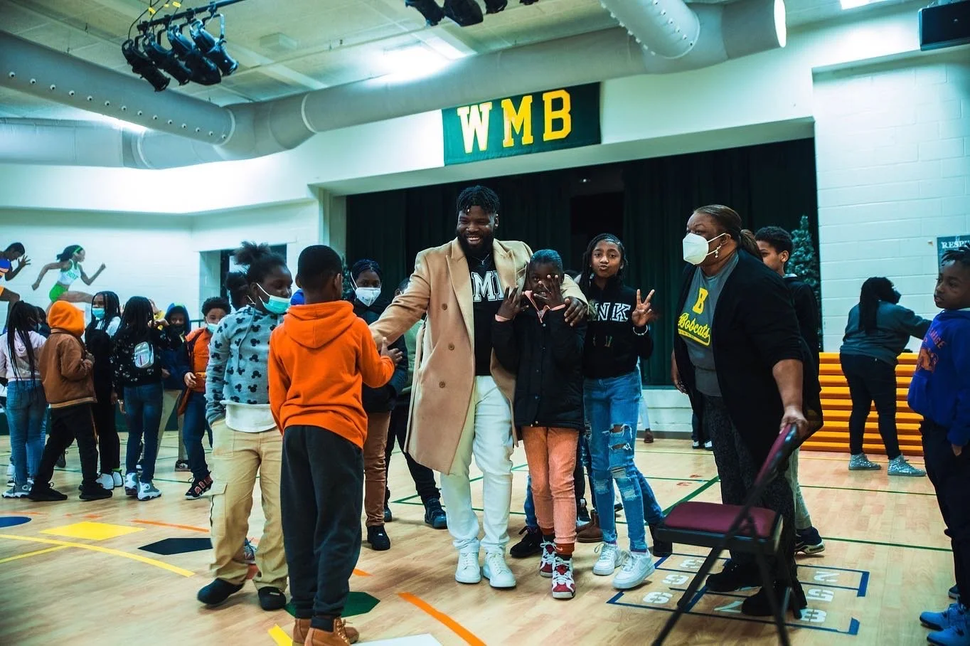 Group of children and adults in a gymnasium, some wearing masks, participating in an event or activity. There is a stage in the background with a green curtain and a sign that reads 'WMB.' People are standing and socializing, with some smiling and posing for the photo.