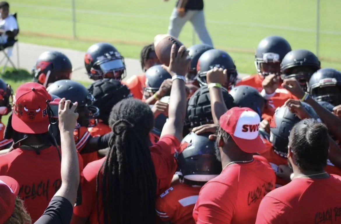 Youth football team in red uniforms and helmets celebrating on the field with one player holding a football up.