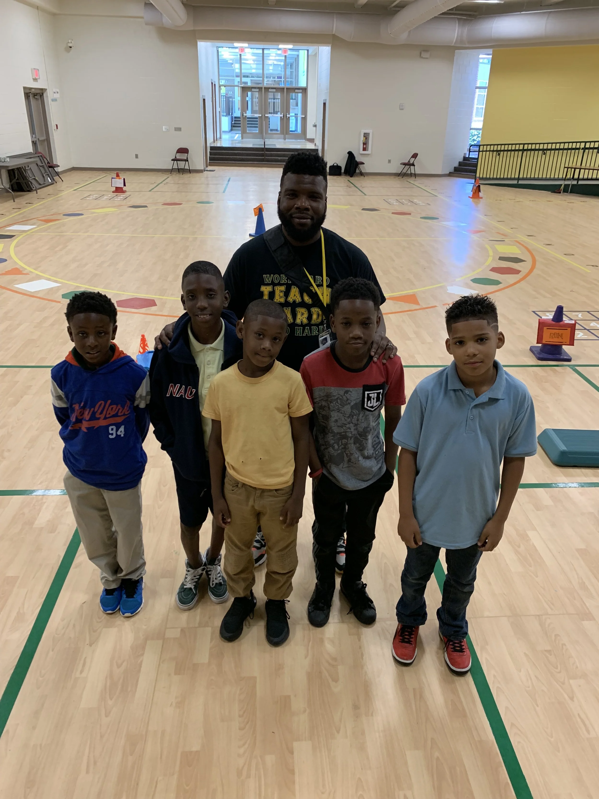 A group of six children and one adult male standing on a gymnasium floor, posing for a photo.
