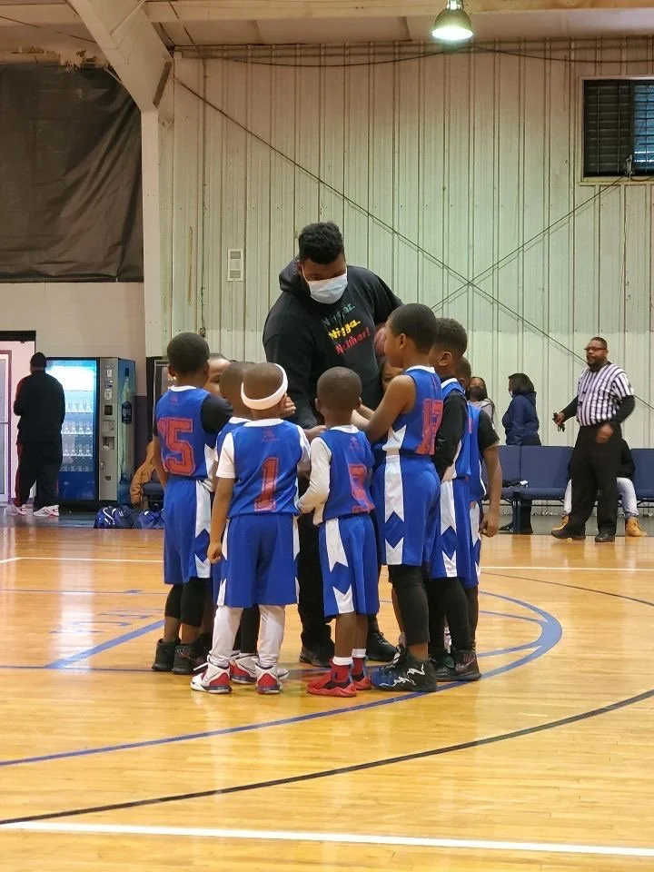 A group of young boys in blue basketball uniforms gathered around their coach, who is wearing a face mask, on an indoor basketball court.