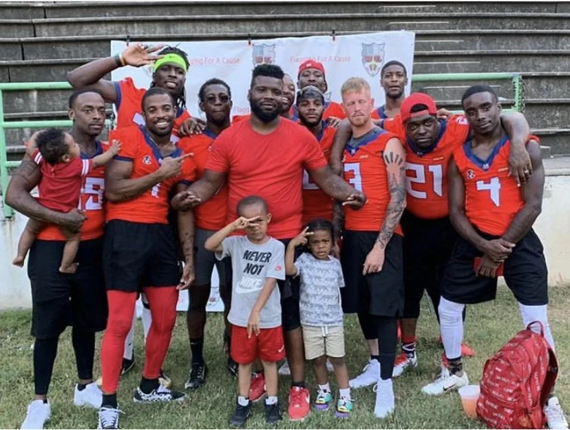 A group of eleven men and two children posing outdoors in front of a sports-related backdrop. Most of the men are wearing red sports jerseys with numbers, and some are making gestures or smiling at the camera.