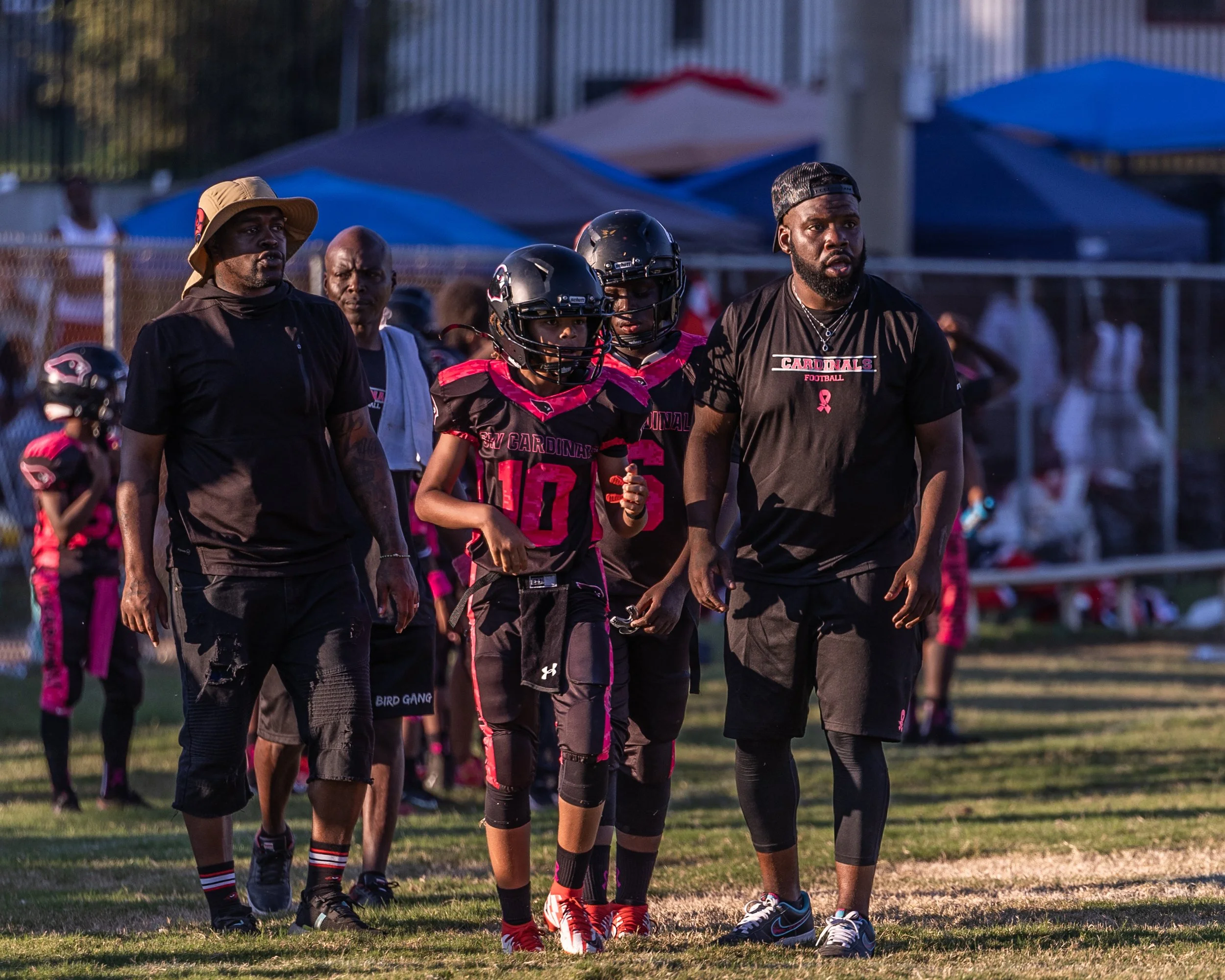 Youth football players and coaches walking on a football field during a game, with tents and spectators in the background.