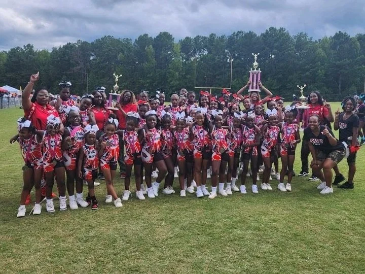 A large group of young cheerleaders in red, black, and white uniforms posing on a football field, holding trophies, with coaches, in an outdoor setting with trees in the background.