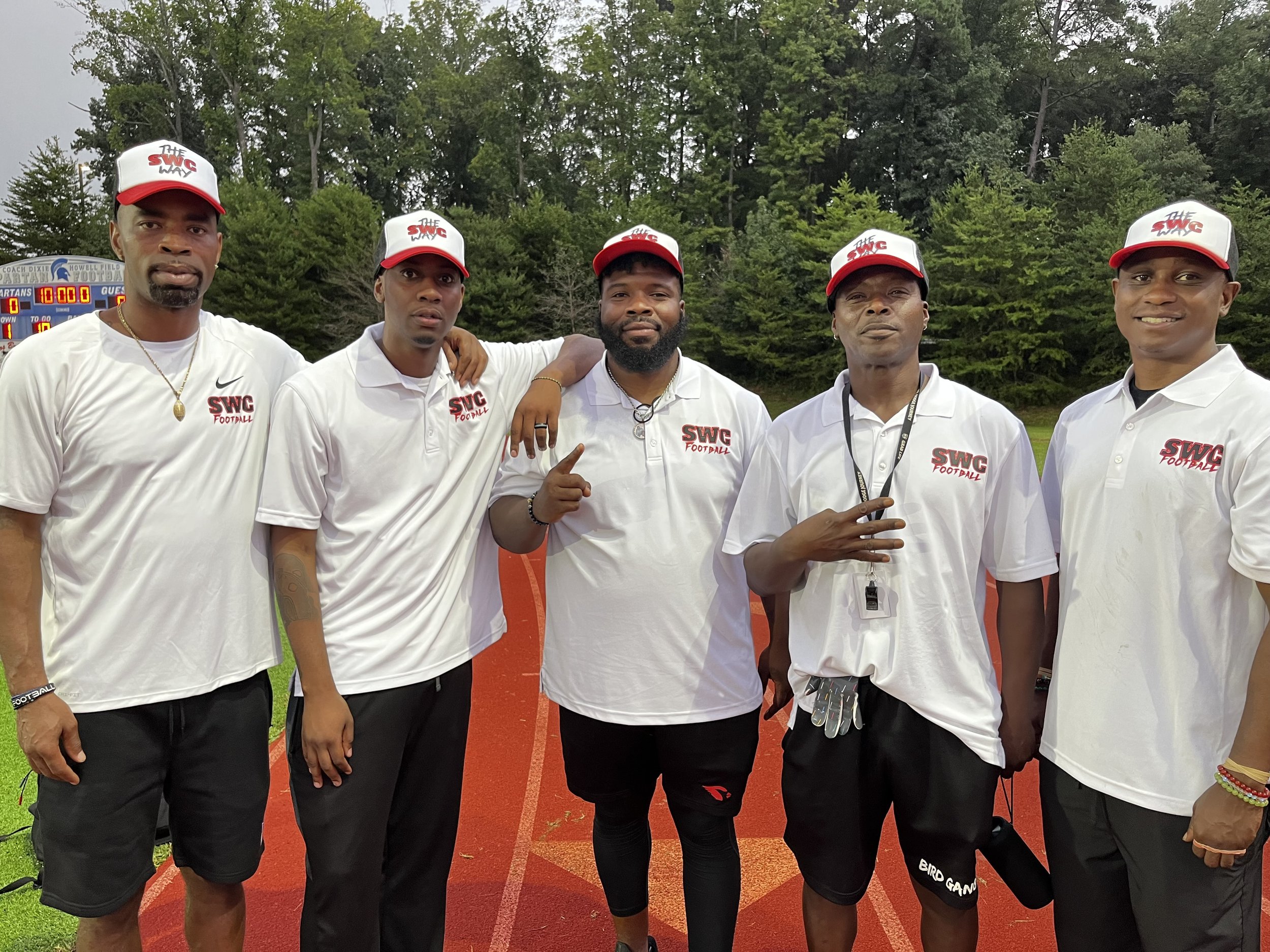 Group of five men in white shirts and caps with 'SWG Football' logo, standing on a running track outdoors with green trees in the background.